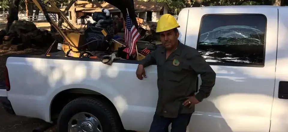 Salim Limon in a hard hat, leaning against his work truck with the bed loaded with cut wood